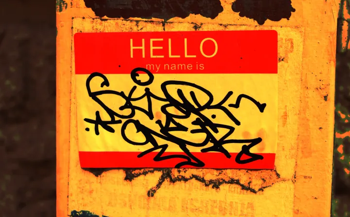 Filtered photo of a "Hello my name is" name-tag sticker on a street post. Over the space for the name is a black graffiti tag.